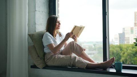 Beautiful girl student is reading interesting book turning pages sitting on window-sill at home in modern apartment. Literature, youth culture and interiors concept.