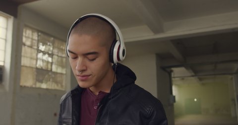Front view of a hip young mixed race man in an empty warehouse, listening to music with headphones on and eyes closed
