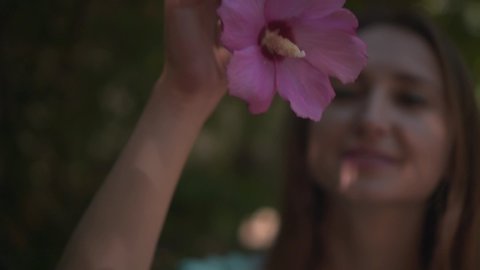 Portrait of a cute woman sniffing and touching the purple hibiscus flowers. Girl in a T-shirt with long hair near blooming hibiscus.