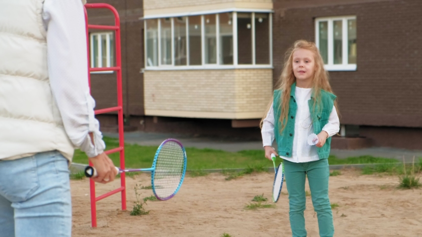Beautiful little preschooler girl with long wavy hair in mint-colored clothes in summer plays badminton with her mother at the children