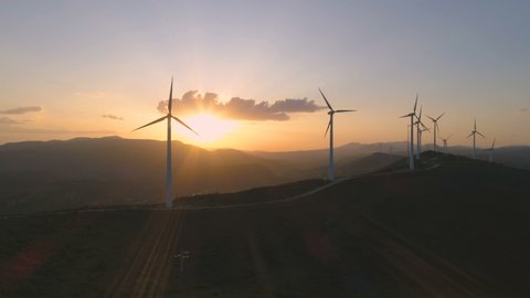 Wind turbine eco farm on beautiful golden hour evening mountain landscape. Renewable energy production for green ecological world. Aerial view of wind mills farm park, evening mountain. Flying forward