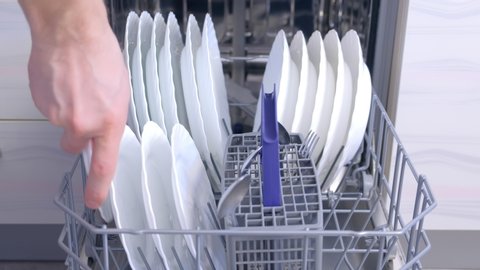 Man loading dishwasher putting tableware and kitchenware in basket, hands closeup. Man putting white utensil and cutlery. washing dishes in kitchen, chores. Household duties using modern appliances.