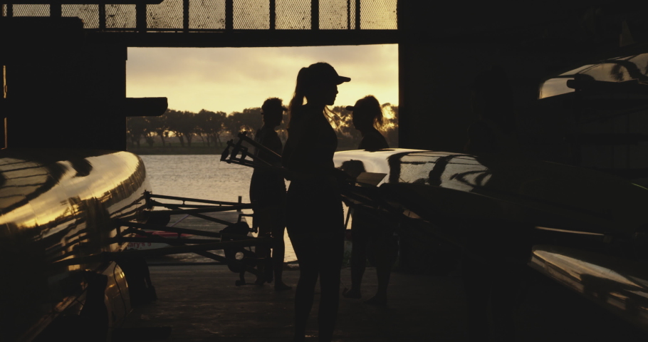 Side view of a team of four young adult Caucasian female rowers on a river lifting a racing shell out of a boatshed before training on a river in slow motion Royalty-Free Stock Footage #1037559434
