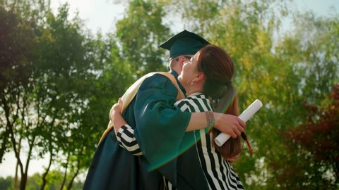 Excited Graduate Student in Gown and Cap with Diploma Hugs his Friend after Graduation Ceremony. 4K Slow Motion Medium Close-Up Shot with Beautiful Sun Lens Flare