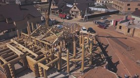 Aerial view of new wooden cottage construction in progress, workers on the top of wooden columns and construction machinery in summer day. Clip. Process of new building construction.
