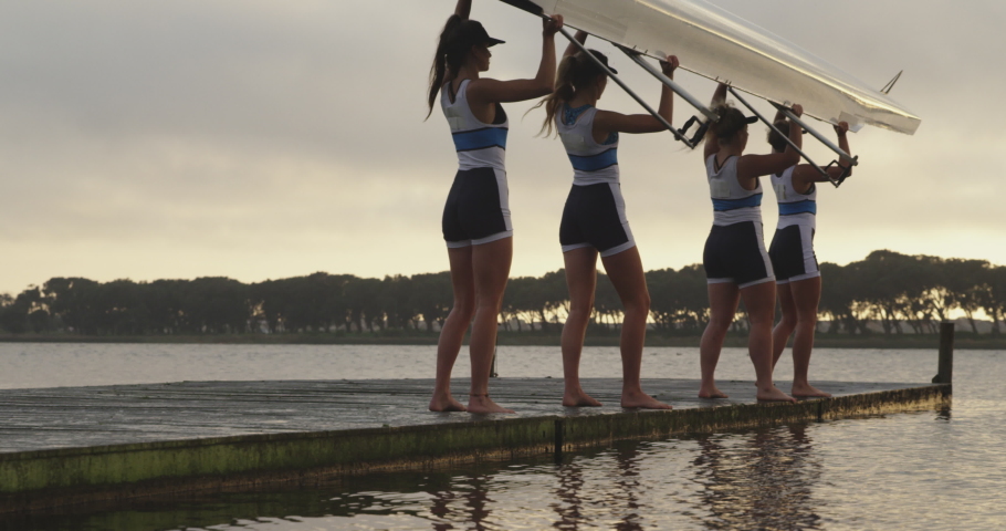 Side view of a team of four young adult Caucasian female rowers standing on a jetty and lowering a racing shell into a river before training in slow motion Royalty-Free Stock Footage #1037568209