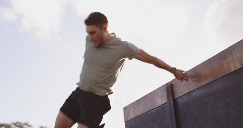 Front view of a young Caucasian man vaulting over a wall at an outdoor gym during a bootcamp training session in slow motion