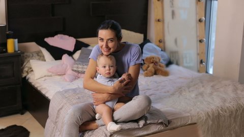 Portrait of a cute young mother hugging her little son in a cozy bedroom. Young mother with her 1 years old little son dressed in pajamas are relaxing in the bed at the weekend together.