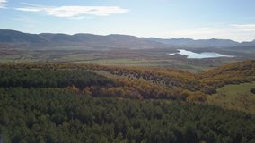 Side flight over Autumn colorful forest and lake surrounded by mountains. 4k aerial view of autumn trees, mountains and lake in Crimea on a sunny day