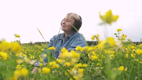 Happy relaxed mature woman sits with her eyes closed wearing headphones and listen music on field, around her wind-moved flowers and insects fly. Sonic Therapy. Sensitivity to nature. Slow motion.