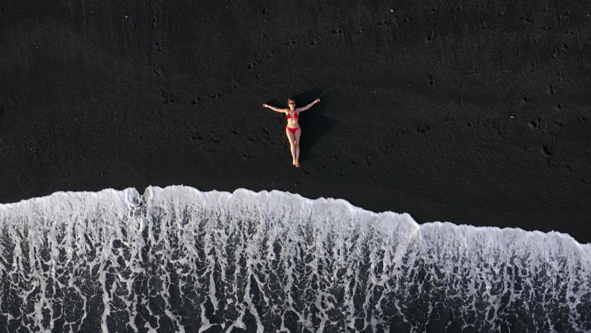 Top view of a girl in a red swimsuit lying on a black beach on the surf line. Coast of the island of Tenerife, Canary Islands, Spain. | Shutterstock HD Video #1037595272