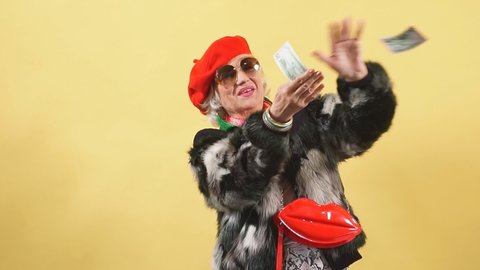 Happy glamour senior old lady in sunglasses throwing stack of dollar bills. Isolated yellow background. Fashion, successful business