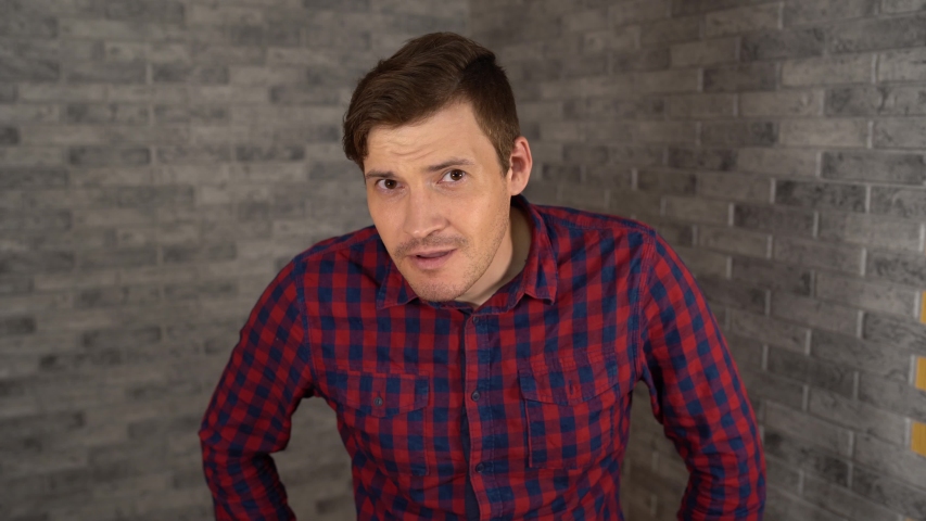 A young handsome man in plaid shirt scratches his head and looks at the camera on a gray brick background.