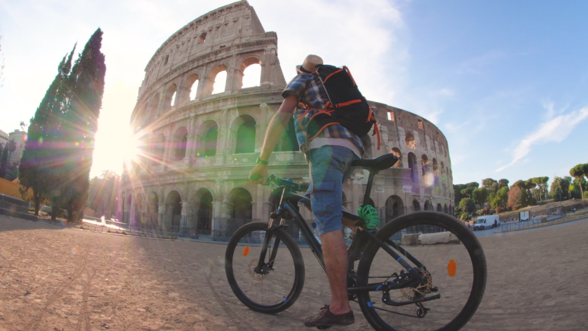 Happy young man tourist riding a bike wearing backpack, shirt and hat at colosseum in Rome, Italy at sunrise.