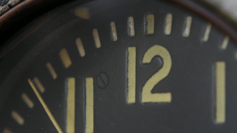 Close-up of the dial of a vintage aviation clock. The seconds are ticking, approaching 12 o'clock.	