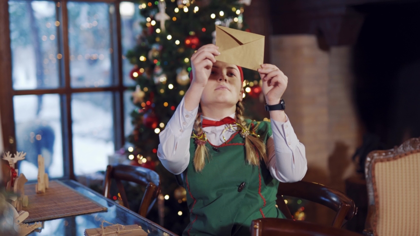 Cute elf with envelope on the background of Christmas lights. Happy female elf looking at envelope attentively while sitting at the table.