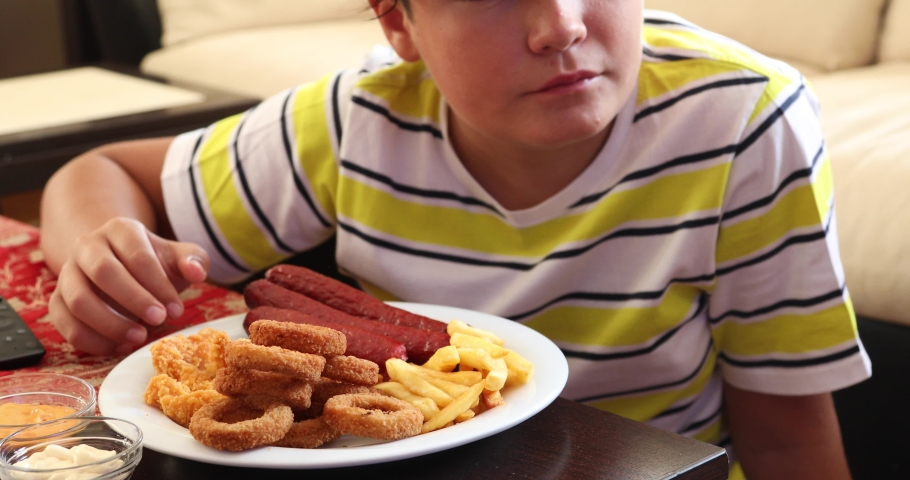 Male teenager with remote controller watching tv and eating fast food at home