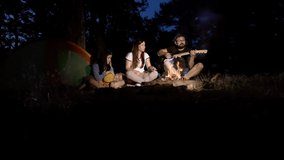 Young men and women are singing songs to the guitar resting around campfire and enjoying music and good company on summer night. The baby is resting next to her mother. Slow motion video.