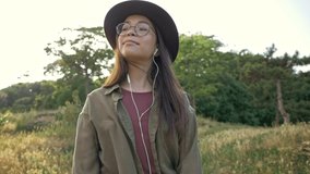 Beautiful smiling young brunette woman in brown hat listening music with earphones and looking around while walking in the park