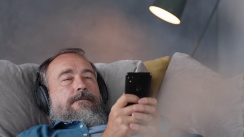 Close up of elderly man with headphones on his head lying on couch and turning audio book on his mobile phone then looking at camera