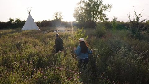 Aerial drone shot of two mature woman walking through countryside to meet friends camping outside teepee - shot in slow motion