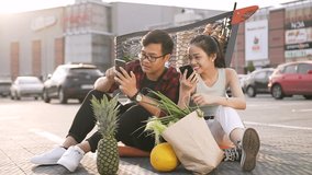 Attractive smiling 30s couple relaxing after shopping,sitting on the ground near big store and watching amusing pictures on their smartphones