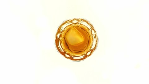Slow motion scene of ice cube dropping into whisky glass isolated on white bright background, top view splashing golden liquor, on the rocks style.