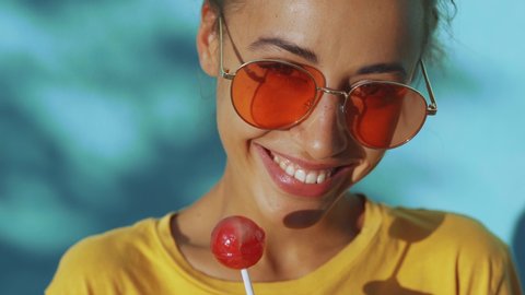 close-up of face of a young tanned emotional woman with big red lollipop. cheerful woman with red eyewear standing against blue background, licking lollipop and showing different emotions.