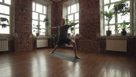 Man practice warrior yoga pose in studio with brick wall. Sporty man in shorts doing yoga indoors with wooden floor and big windows with copyspace. Wide angle