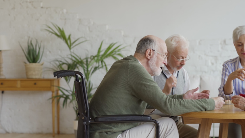 Tracking shot of group of four cheerful retired senior people, two men and two women, having fun sitting at table and playing bingo game together in nursing home Royalty-Free Stock Footage #1037667410
