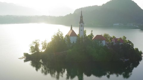 Lake Bled at sunrise with mild lens flare lighting the Church island of the assumption of Mary, Aerial footage on a beautiful Slovenian Morning.