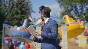 Young caucasian businessman in suit stands in the middle of amusement park and discusses project during video call using touchpad. Doing business everywhere