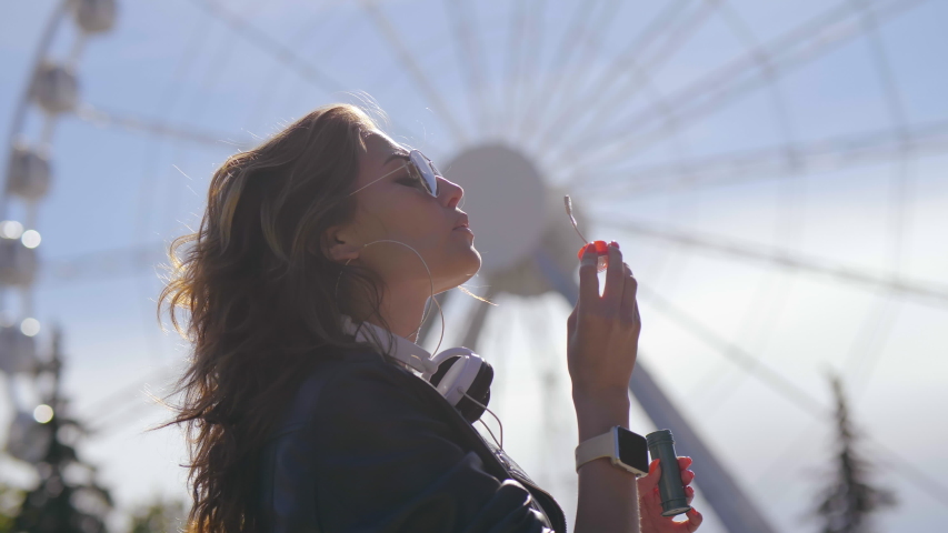 Gorgeous caucasian girl blows soap bubbles on background of ferris wheel in amusement park. Close up side view