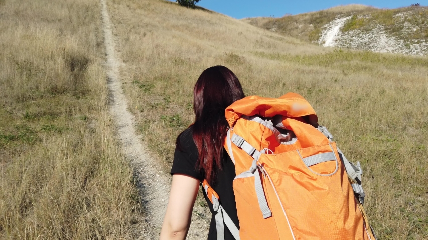 A young woman in black clothes with a large orange backpack on her back in her hands is climbing the mountain.
