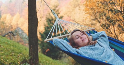 Woman Wakes Up after Sleep in a Hammock in Autumn. SLOW MOTION. Young woman daydreams, unwinds in a calm fall outdoor, rural country nature with colourful forest in background. Cozy season.