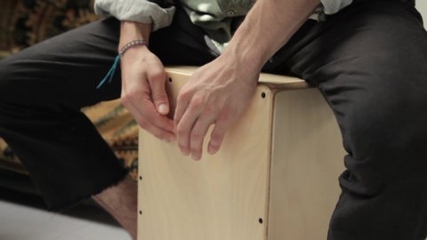 Detail shot of a young gypsy styled man hands playing cajon flamenco drumbox having fun rehearsing and creating percussion sounds on a home studio background. Rythm and compass concept.