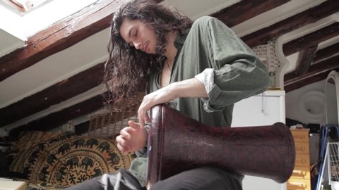 Medium down shot of a young handsome long haired man having fun learning playing arabic darbuka having fun rehearsing and creating percussion sounds on an alternative exotic home studio background.
