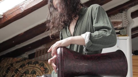 Medium down shot of a young handsome long haired gypsy styled man focused playing arabic darbuka having fun rehearsing and creating percussion sounds on an alternative exotic home studio background.