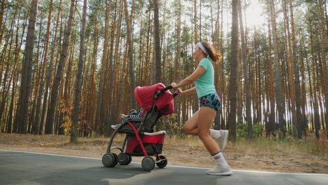 Running American girl with baby stroller enjoying a sunny summer day in a pine forest. Jogging or power walking, active family with baby jogger. Slow-motion 4k video