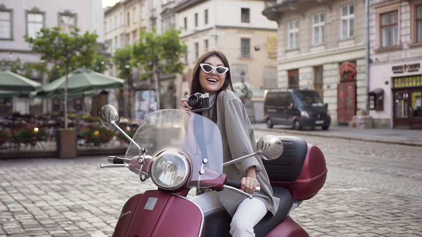 Good-looking happy 30s girl in fashion sunglasses keeping outmoded camera sitting on the cool scooter on the background wonderful architecture