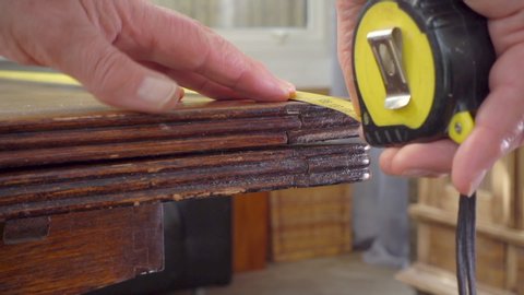 Close POV shot of a man’s hands slowly pulling a traditional retracting steel tape measure across the length of an old wooden table, then checking the measurement.