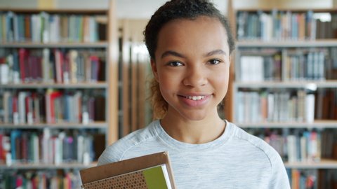 Portrait of attractive African American girl student holding books in high school library smiling looking at camera. Education, literature and people concept.