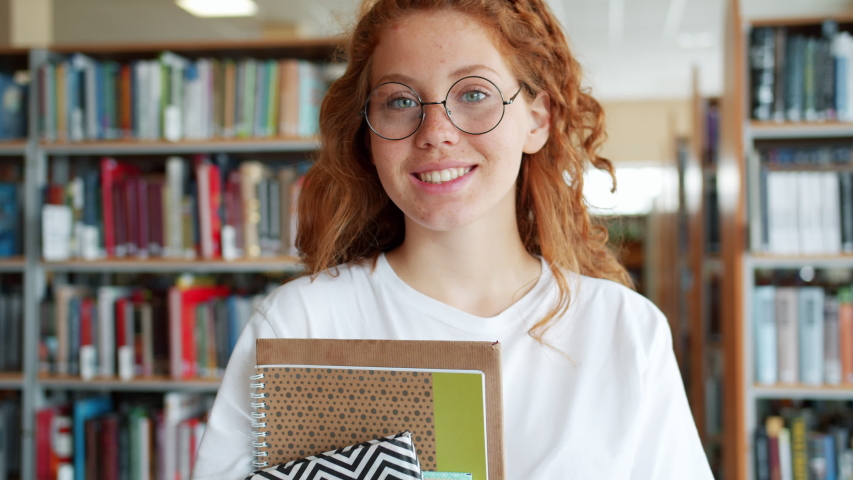 Portrait of joyful student pretty girl holding books in university library smiling looking at camera indoors alone. People, education and emotions concept.