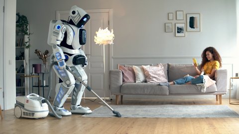 A cyborg is hoovering a room with a lady in it