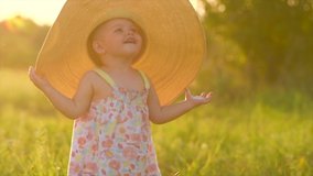 Adorable little baby girl on a field, enjoying nature outdoors wearing wide brimmed straw hat. Beautiful cute little funny one year old child smiling and doing her first steps. Slow motion 4K UHD.