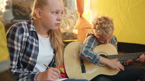 Teenager girl writing song to guitar music in camping tent. Boy teenager playing music on acoustic guitar in tourist tent. Brother and sister spending leisure together in summer hike