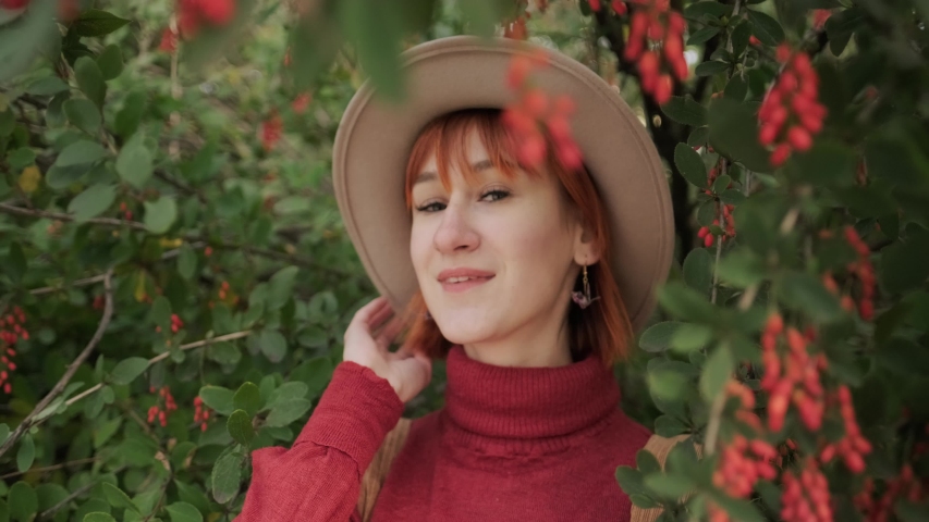 Young attractive red-haired girl with a short hair in a hat and a teracotta sweater in a natural park on an autumn day. Barberry tree with red berries on a background.