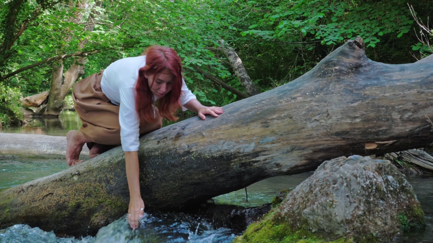 A red-haired girl dressed in a rustic style drinks clean river water and washes her hands.