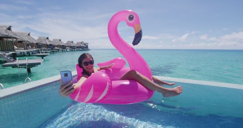 Vacation travel woman on inflatable Flamingo float mattress using mobile cell phone taking selfie in swimming pool. Girl relaxing sunbathing enjoying holidays resort pool in bikini. Luxury lifestyle.
