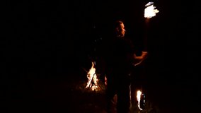 Low key. Young male with long hair rotates burning torch outdoors on a black night video slow motion. Modern fakir does tricks with a burning staff
